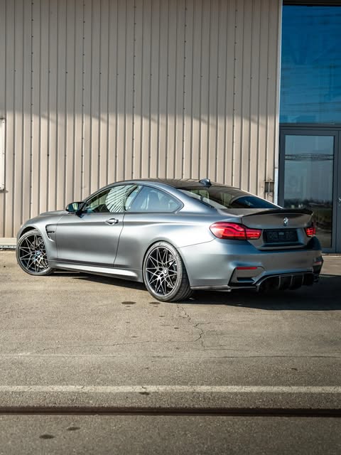 BMW M4 Competition in Mattgrau, Heckansicht von schräg hinten vor einer Industriehalle